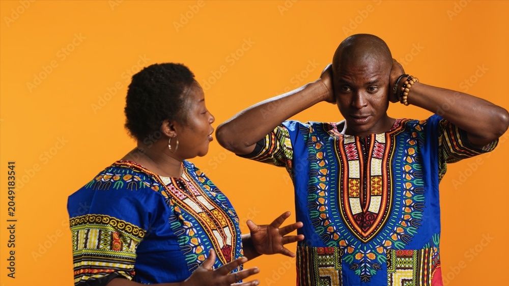 Obraz premium Furious girlfriend shouting at her boyfriend and fighting, having disagreement over orange background. Frustrated man covering his ears and trying to ignore his wife yelling in studio.