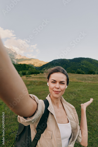 Woman takes a casual outdoor selfie in a green countryside field with a backpack  smiling under a bright sky with distant hills during a hike beside nature