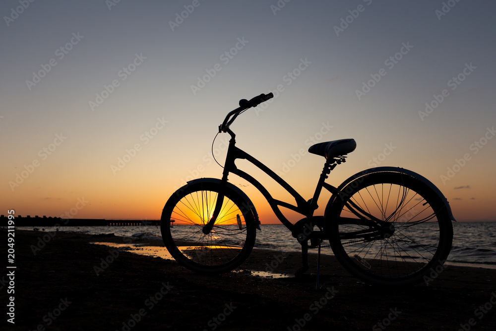 Fototapeta premium Minimalist shot of a vintage cruiser bicycle silhouette standing on a sandy shore against a dramatic orange sunset sky and calm sea waves.