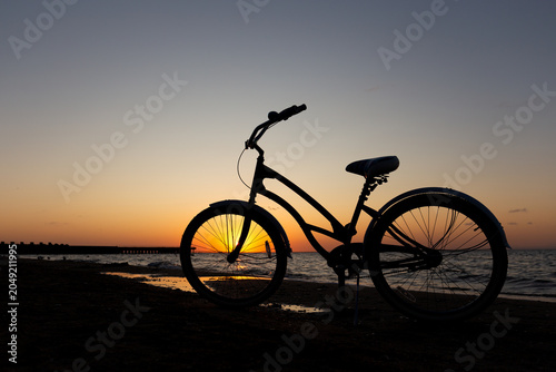 Fotografi Minimalist shot of a vintage cruiser bicycle silhouette standing on a sandy shore against a dramatic orange sunset sky and calm sea waves