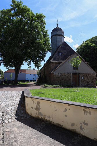 Historische Dorfkirche mit markantem Turm hinter einer Mauer im Sonnenschein, Meuschau, Merseburg, Saalekreis, Sachsen-Anhalt, Deutschland