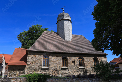 Historische Dorfkirche mit markantem Turm hinter einer Mauer im Sonnenschein, Meuschau, Merseburg, Saalekreis, Sachsen-Anhalt, Deutschland