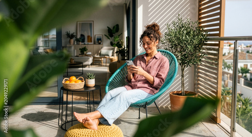 Smiling Young Woman Relaxing Barefoot with Smartphone in Comfortable Chair on Sunlit Modern Balcony with City View