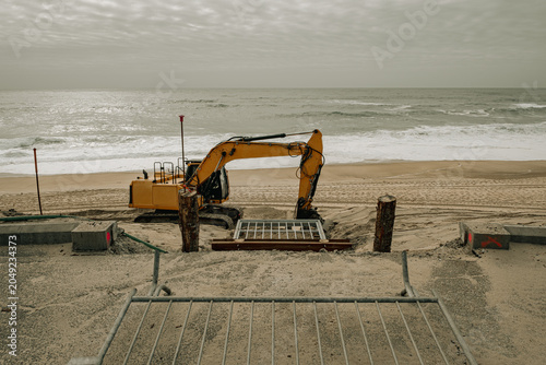 Professional full color photograph of a yellow crawler excavator executing sand dune reconstruction on a beach affected by global warming, climate erosion, and rising sea levels.