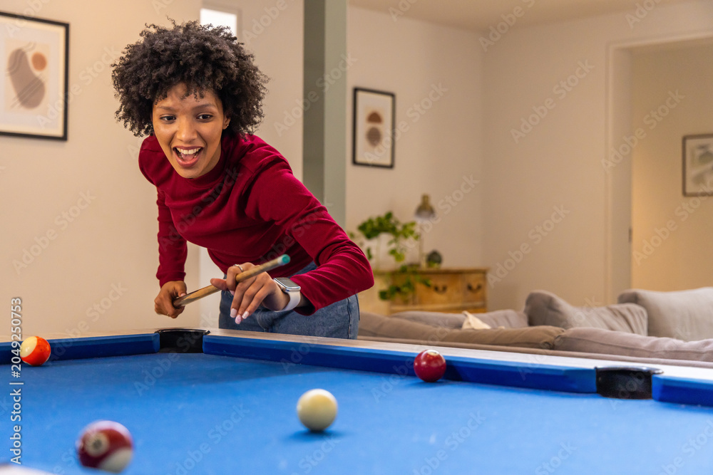 Fototapeta premium African American woman wearing red top leaning over blue-felt pool table at home, holding cue
