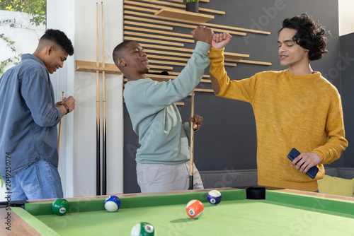 Diverse male teens celebrating by fist bumping near pool table with cues and smartphone