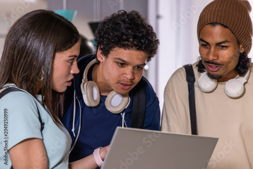 Diverse teen friends leaning over open laptop at school hall, wearing headphones, backpacks, beanie