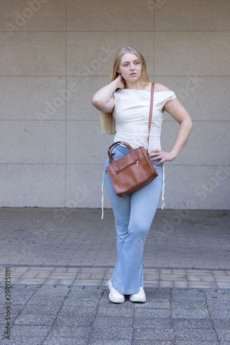 Woman standing at plaza touching hair, wearing off-shoulder top, flared jeans, brown crossbody bag