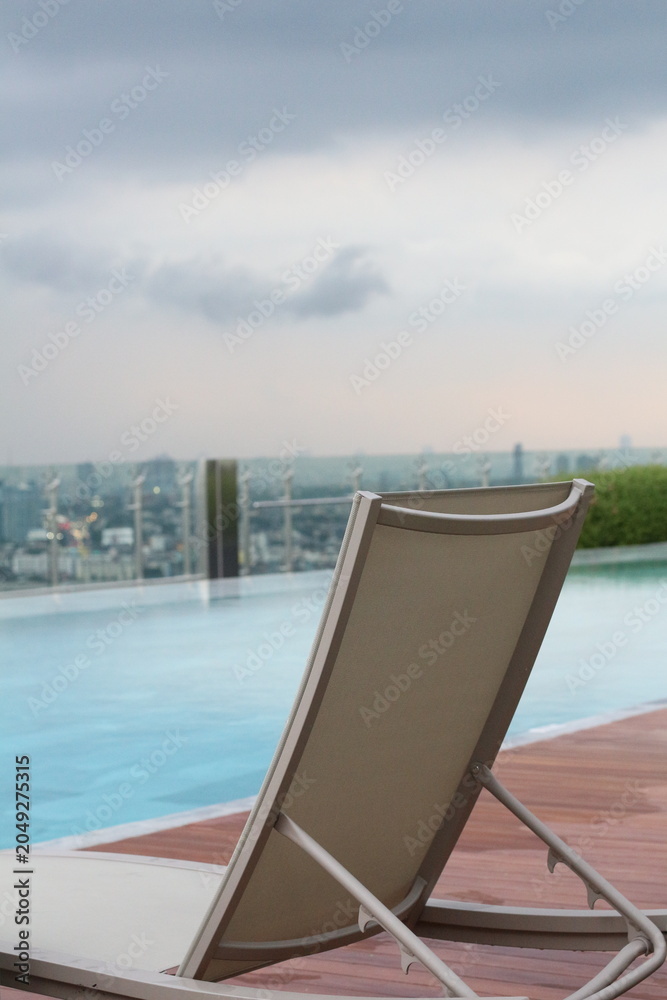 Fototapeta premium Relaxing lounger by infinity pool overlooking city skyline on a cloudy day