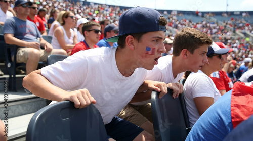 Young male supporter in backward cap, with American flag face paint leaning forward and shouting from seat, outdoor stadium stands, sports spectator lifestyle photography