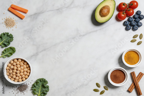 Healthy food ingredients including avocado, tomatoes, spices, seeds, kale, and soybeans on marble background