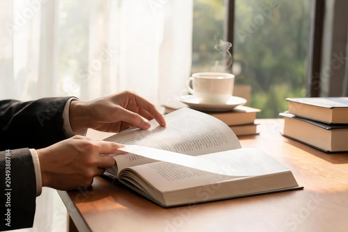 Person Turning Pages of Open Book with Steaming Cup and Stacked Books on Wooden Table by Window