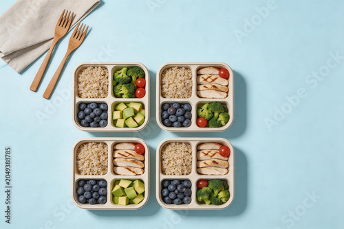 Healthy meal prep containers with grilled chicken, broccoli, cherry tomatoes, blueberries, avocado, and grains on a blue background