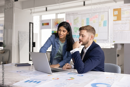 Canvas-taulu Young 30s Indian woman showing presentation on laptop to man colleague or consulting company client during formal meeting in modern conference room