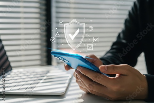 Person holding smartphone with security icons and laptop on desk in office setting with window blinds