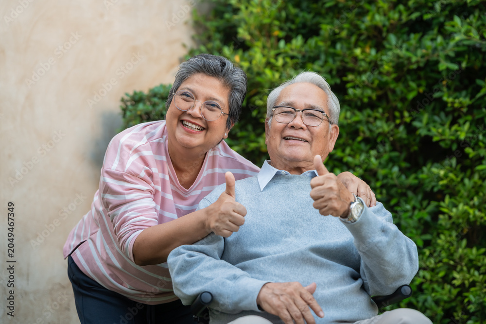 Fototapeta premium Two elderly people sit close together. Woman leans toward seated man. Both give thumbs up. Background shows greenery and a wall. They look happy