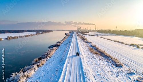 Aerial view of a snowy road flanked by a river and snow-covered land, under a bright sky