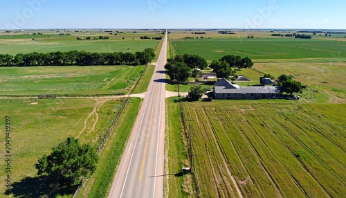 Aerial view of a straight road receding into the distance, flanked by vast fields and farmland under a bright blue sky