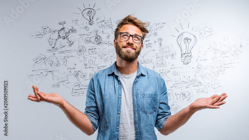 Man with glasses and beard stands in front of whiteboard filled with creative sketches and ideas, looking thoughtful and inspired