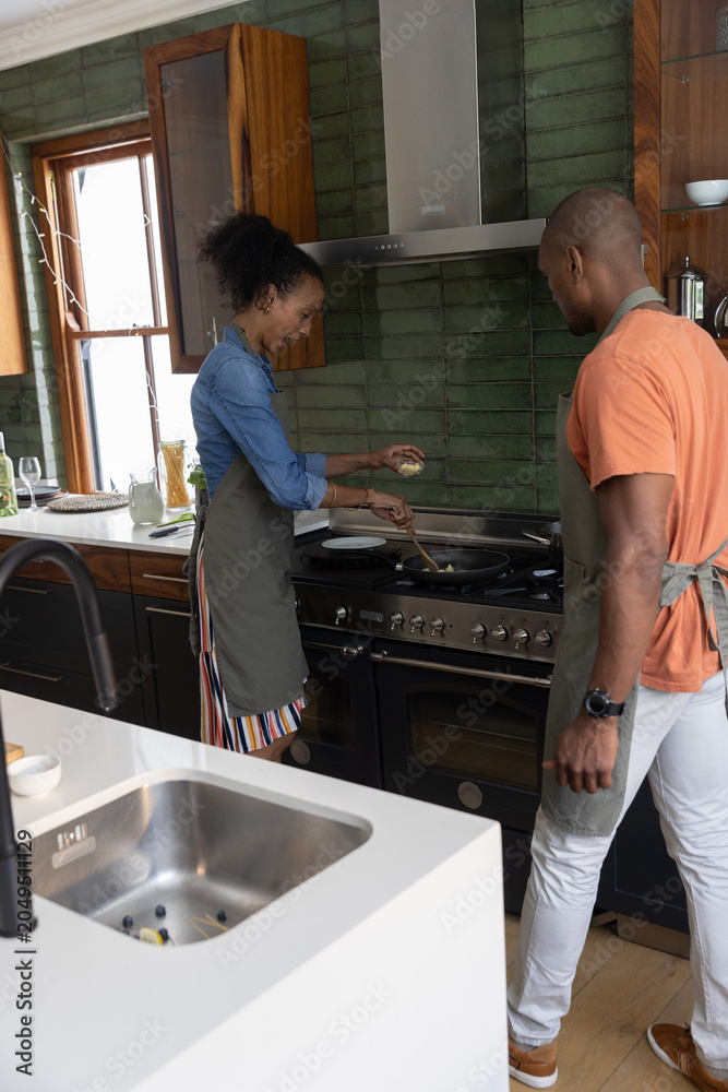 Obraz premium Diverse couple cooking at gas range in kitchen, wearing aprons and stirring food with wooden spoon