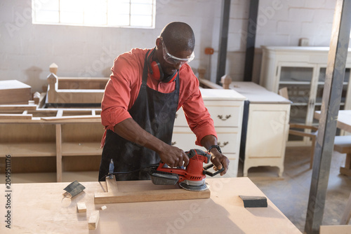 Carpenter sanding wooden plank with red orbital sander at workshop workbench under natural light