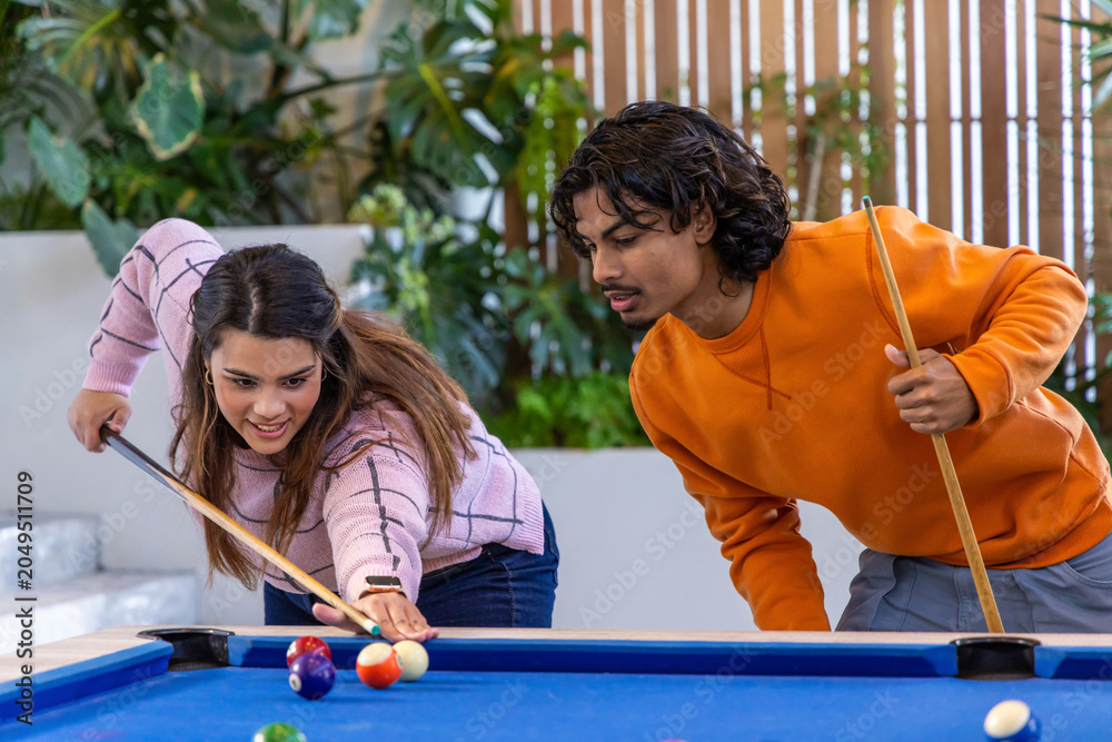 Naklejka premium Two friends leaning over blue-felt pool table in plant-filled lounge, lining up shot with cues