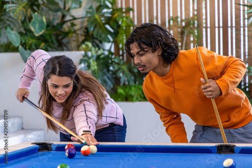 Two friends leaning over blue-felt pool table in plant-filled lounge, lining up shot with cues