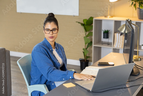 Woman wearing blue denim shirt and glasses, sitting at desk in home office, using laptop, notebook