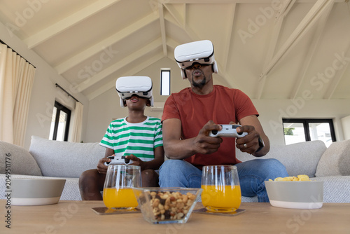 African-American father-son gaming at home wearing VR gear holding pads by table with juice, snack