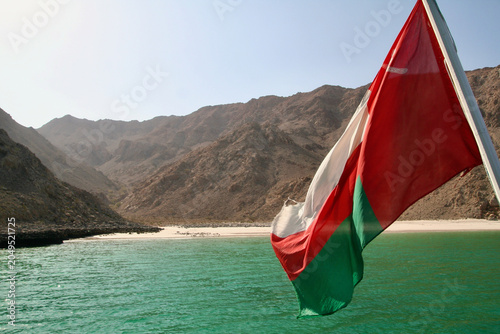 Canvastavla Omani flag flying on dhow boat with turquoise fjord and Hajar Mountains, Musanda