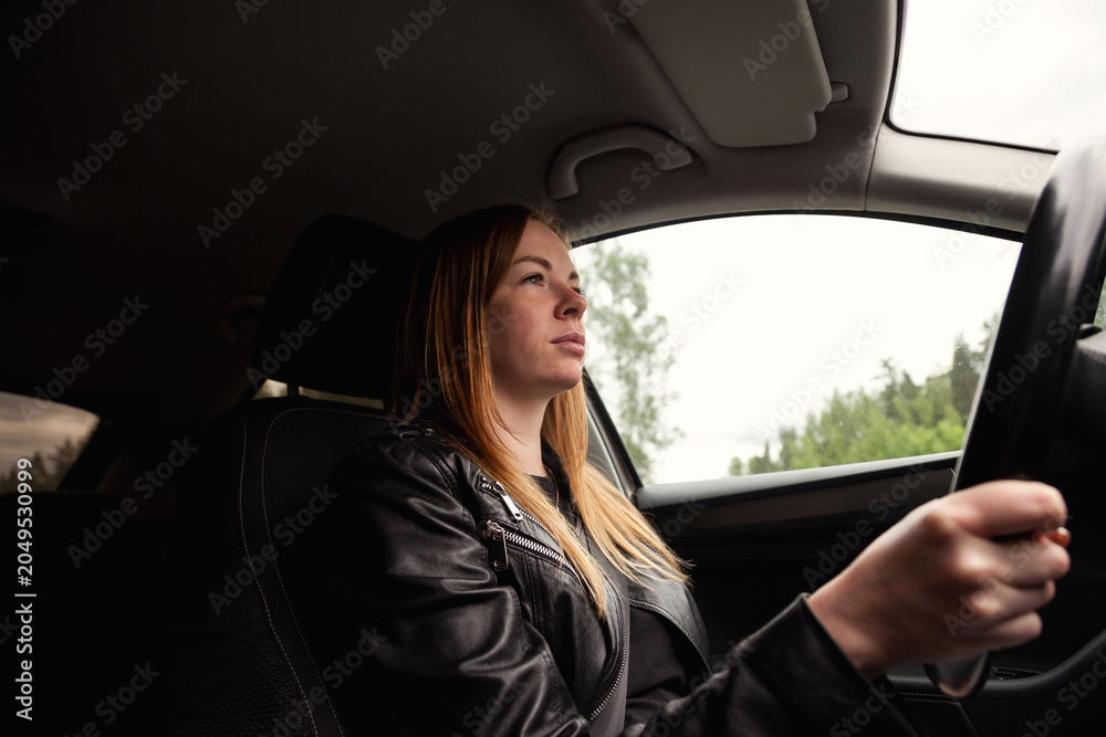 Fototapeta premium A woman is actively driving a car, displaying focus and determination. The lush greenery outside the vehicle contrasts with her leather attire, showcasing a serene afternoon expedition into nature.