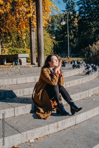 Woman sits on stone park steps surrounded by birds in sunlight. Mindful walking outdoor therapy, slow living autumn routine, connecting with nature footprints.