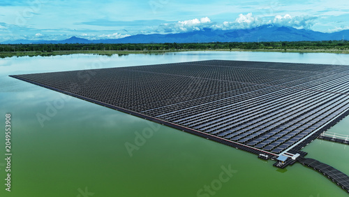 Aerial view of a massive sprawling floating solar farm on a green water reservoir with mountains under a cloudy sky.