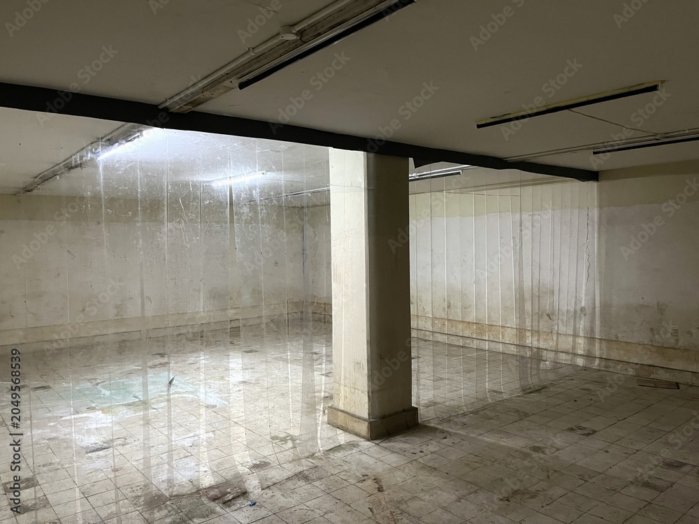 Naklejka premium Spacious underground facility featuring concrete support columns, tiled flooring, and fluorescent illumination within an aging building structure.