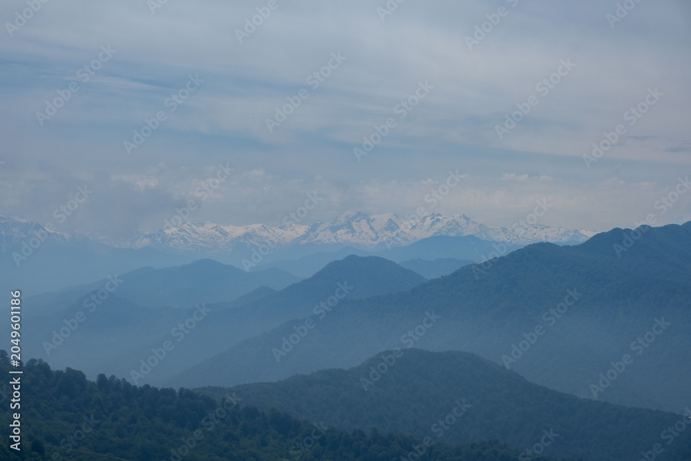 Fototapeta premium Distant snow-capped Caucasus mountain peaks behind misty blue layers of hills under a cloudy sky