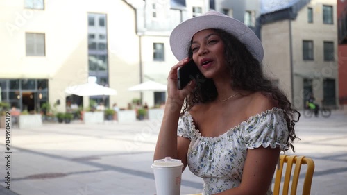 Attractive smiling woman in hat talking on cellphone while sitting in cafe outdoors.