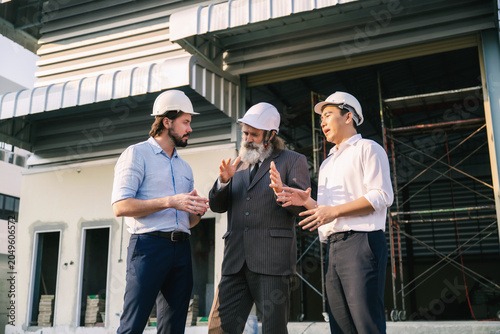 Investor engineers talking outside empty factory warehouse construction site, Three people in white hard hats converse near a building under construction, Customer service or investors planning.