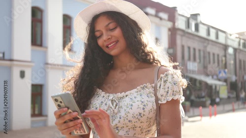 Beautiful young dark curly haired girl using mobile phone while sitting outdoors.
