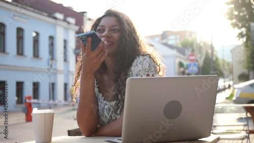 Attractive smiling woman recording voice message on cellphone while sitting in cafe outdoors.