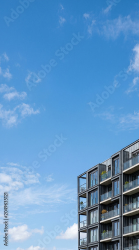 Minimalist architectural detail of a modern apartment building with dark facade and glass balconies, set against a vast blue sky with ample copy space for text.