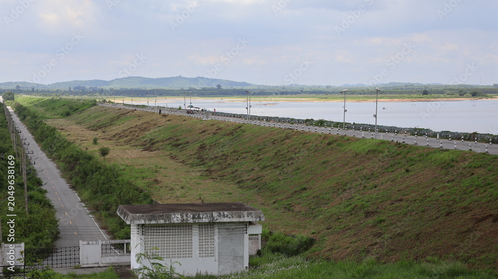 Naklejka premium The image depicts a wide landscape featuring a sloped embankment running parallel to a water body, with a road lined with streetlights on the top edge of the embankment