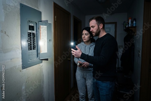 Couple inspecting an electrical breaker panel during a power outage at home, using smartphone and flashlight to troubleshoot household electricity issues in a dark residential hallway.