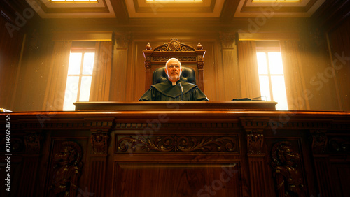 Senior male judge sitting at a high wooden bench in a formal courtroom. The official wearing a black robe presides over a trial in dramatic lighting. Law, justice, and authority concept for legal.