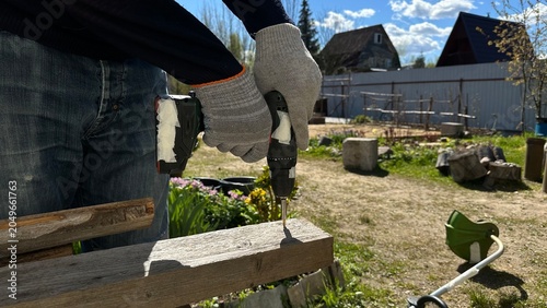 Hands in protective work gloves using a power drill on a wooden surface during home renovation. DIY woodworking, cottage repair, construction, craftsmanship and industrial work.