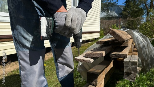 Hands in protective work gloves using a power drill on a wooden surface during home renovation. DIY woodworking, cottage repair, construction, craftsmanship and industrial work.