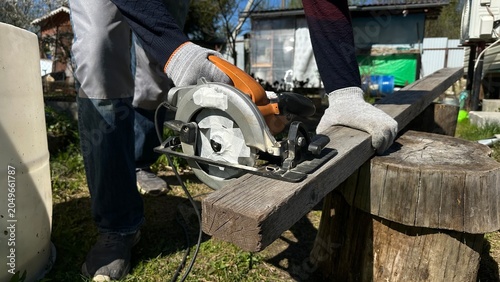 Hands in protective work gloves using a circular saw on a wooden surface during home renovation. DIY woodworking, cottage repair, construction, craftsmanship and industrial work.