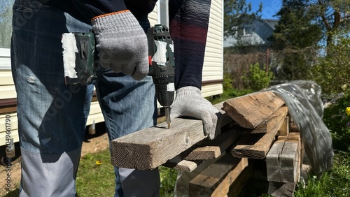 Hands in protective work gloves using a power drill on a wooden surface during home renovation. DIY woodworking, cottage repair, construction, craftsmanship and industrial work.