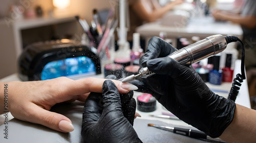 Professional manicurist in black gloves using an electric nail drill for hardware manicure treatment on client's fingernails in modern beauty salon with UV lamp and polish bottles background.