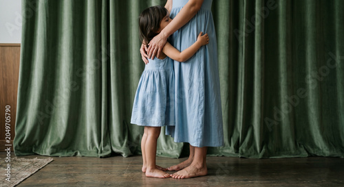 Tender mother and daughter embracing indoors while wearing matching blue dresses. Intimate family moment expressing love, security, emotional connection, parenting