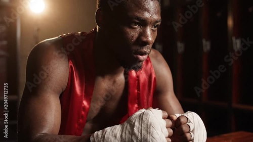 Sweaty exhausted boxer in red robe and hand wraps resting in a dark locker room after an intense workout, focused expression